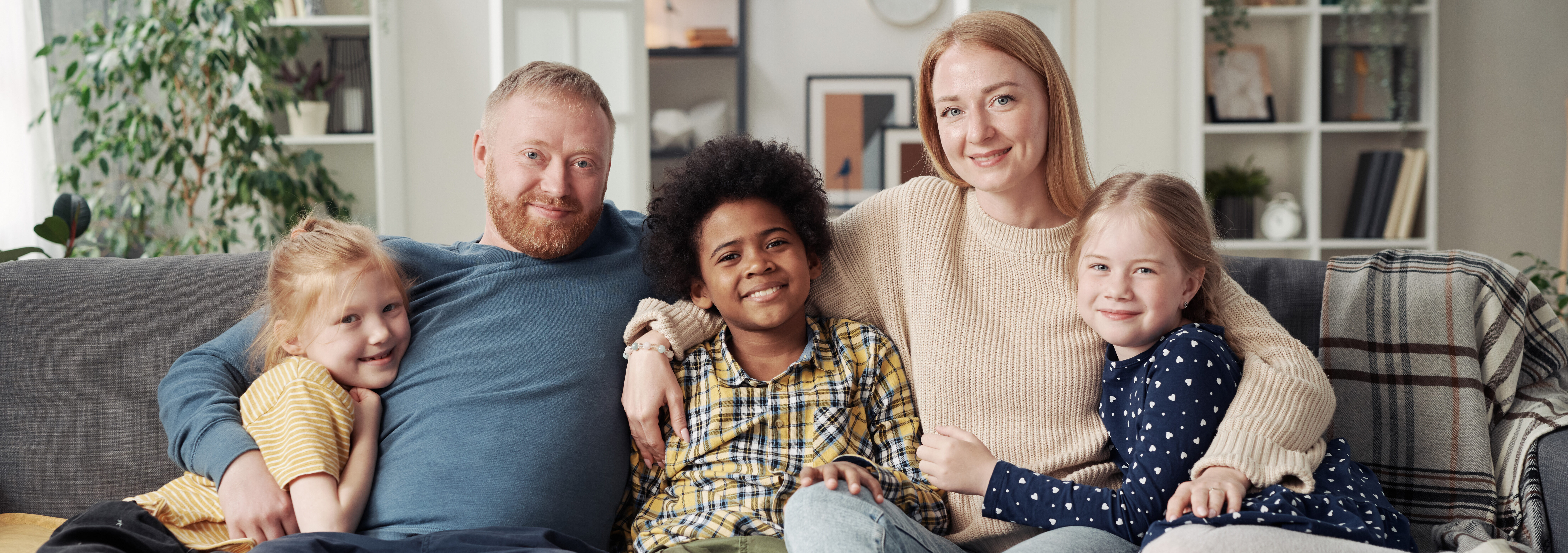 Portrait of happy adoptive family with their children embracing together and smiling at camera while resting on sofa in living room